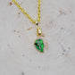 A gold chain necklace with a small leaf-shaped pendant featuring green enamel and white leaf veins, displayed on a light wood slice background.