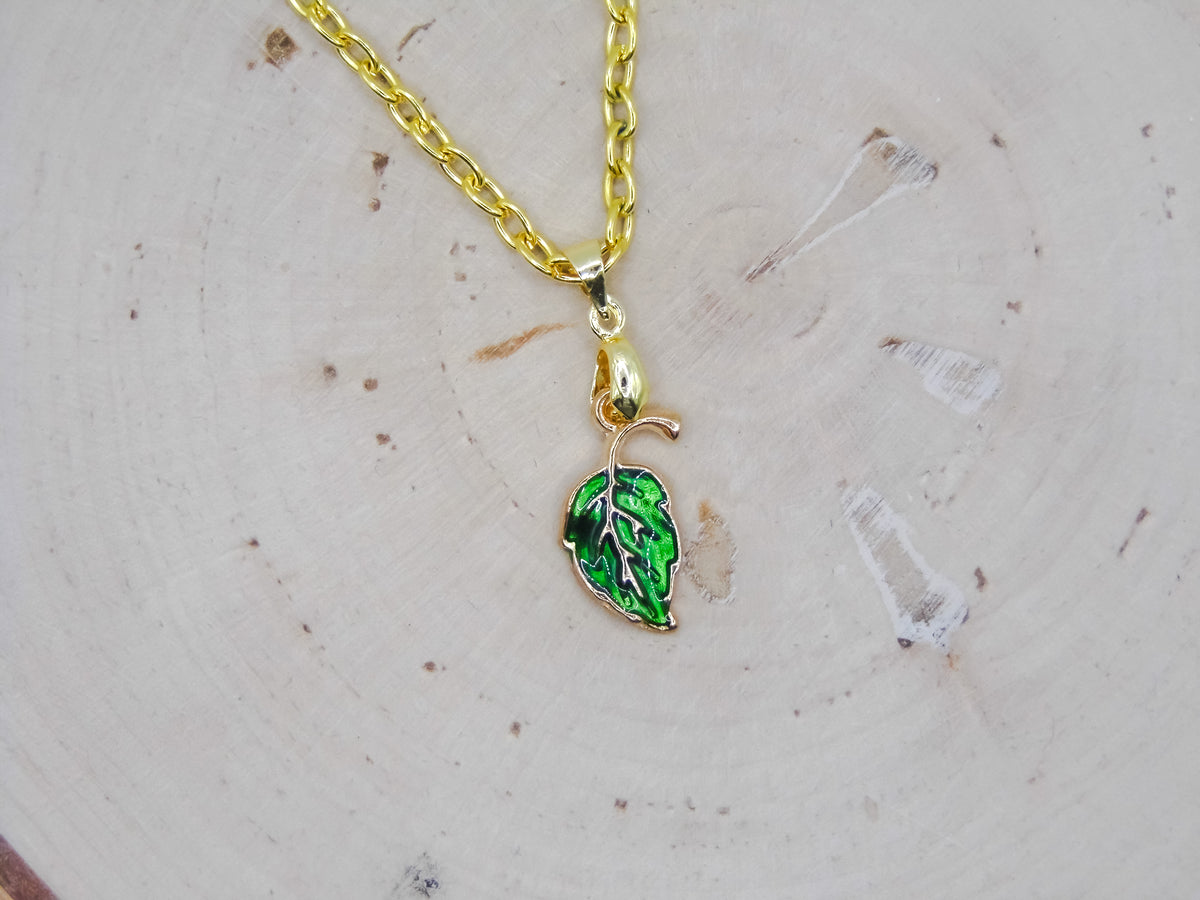 A gold chain necklace with a small leaf-shaped pendant featuring green enamel and white leaf veins, displayed on a light wood slice background.