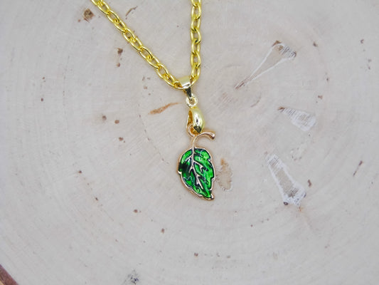 A gold chain necklace with a small leaf-shaped pendant featuring green enamel and white leaf veins, displayed on a light wood slice background.